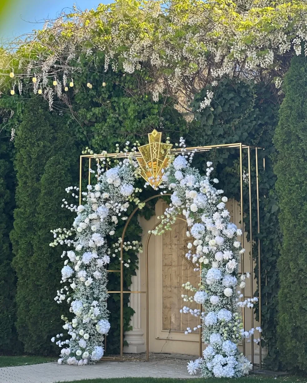 Arcadă aurie art-deco cu flori albe, decor intrare eveniment printre tufe verzi